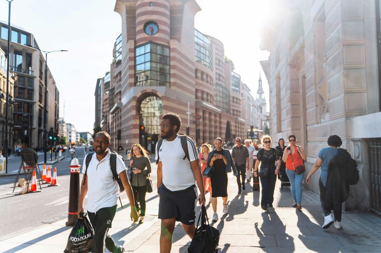 Pedestrians walk along a sunny city sidewalk lined with tall buildings