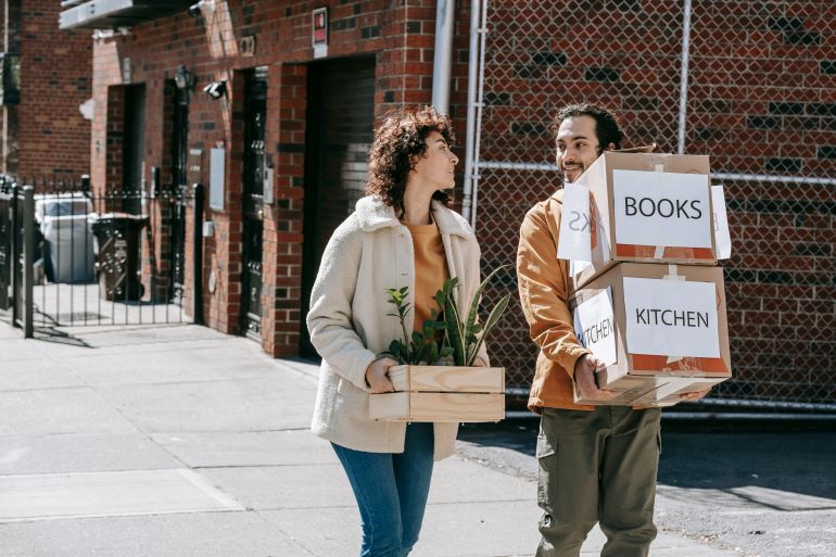 Two people carry moving boxes and plants on a city sidewalk, showing a neighborhood move into an urban residential area.